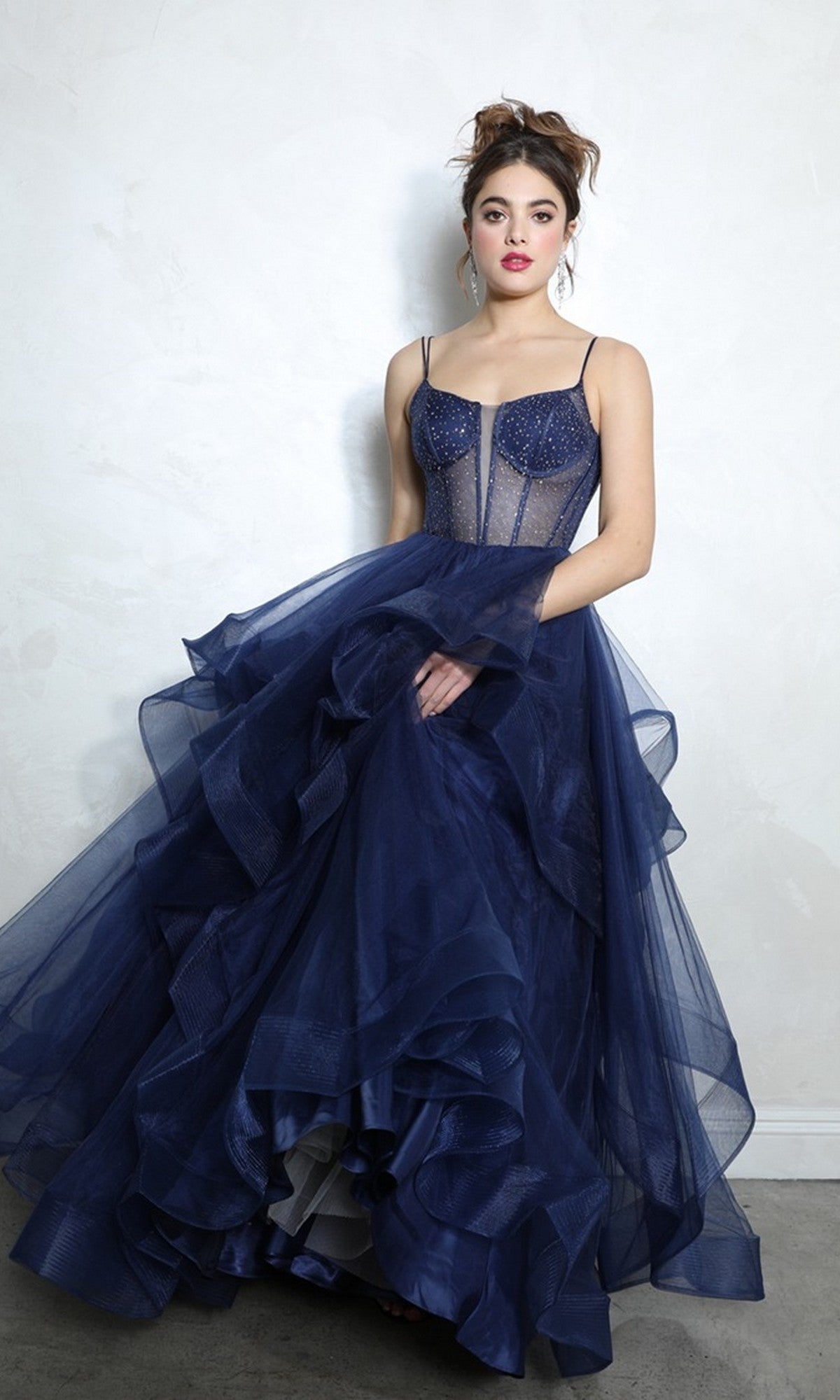 A woman in the Eureka 9950 long prom dress in navy, featuring a beaded corset bodice and voluminous layered tulle skirt, stands against a plain light-colored wall.